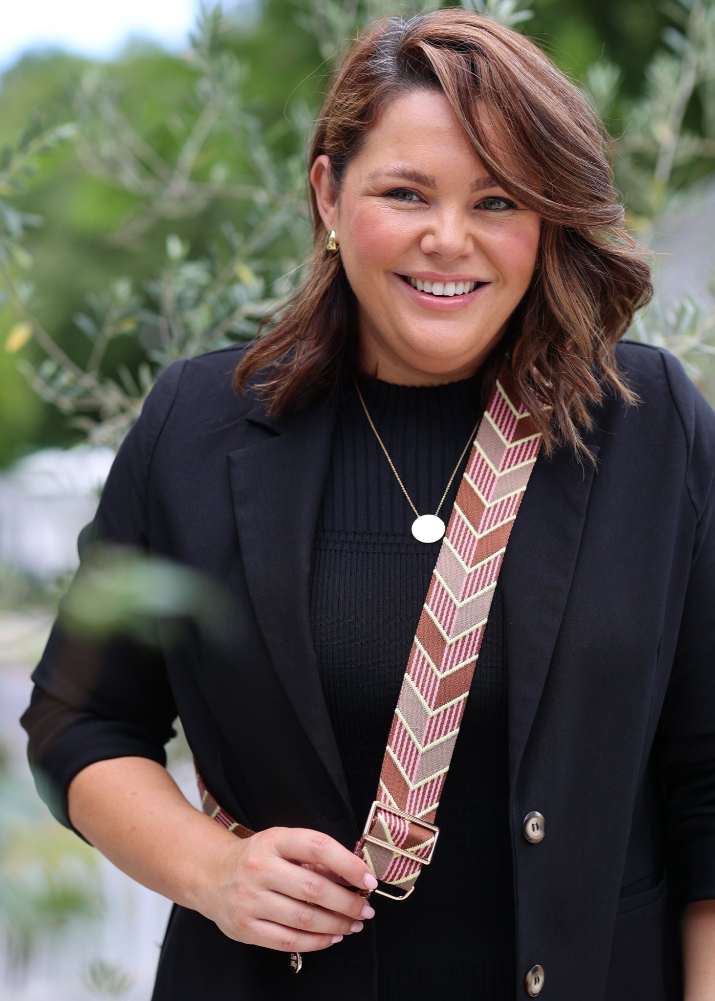 A person wearing a black jacket and a smiling while showcasing a brown/yellow/pink arrow-patterned guitar strap over their shoulder.