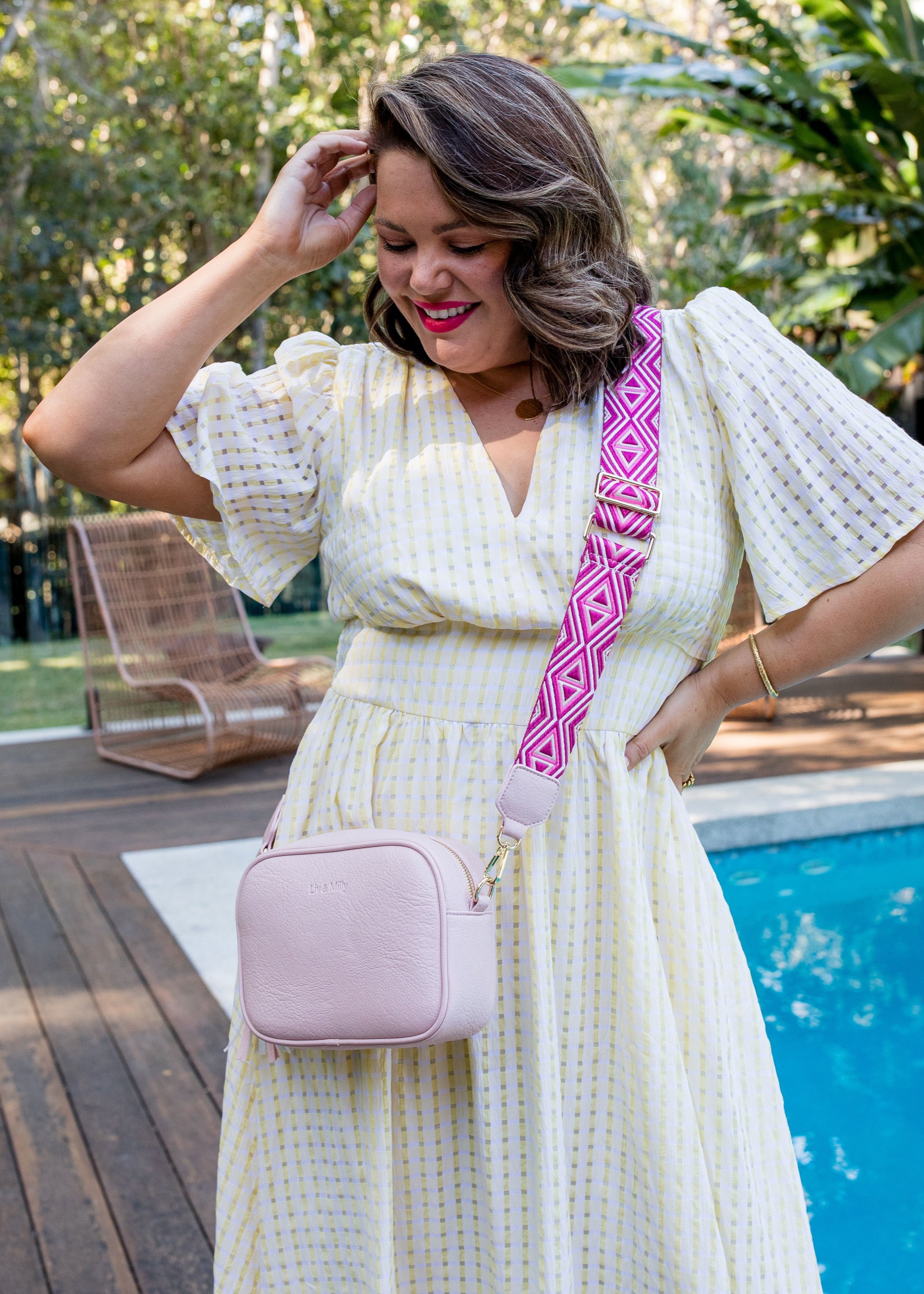 A person wearing a pastel pink crossbody bag with a vegan leather finish, gold hardware, and an adjustable strap. The bag is shown in a casual outdoor setting near a pool.